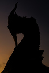 Silhouette of Thai Naka, big snake, serpent statue on yellow  sky at dawn. Mythical creature in Buddhism belief. Traditional Thai style architecture.