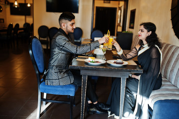 Lovely indian couple in love, wear at saree and elegant suit, sitting on restaurant and cheers together by orange juice.