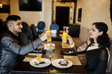 Lovely indian couple in love, wear at saree and elegant suit, sitting on restaurant and cheers together by orange juice.