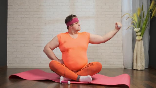 Funny fatso is engaged in fitness using dumbbells sitting on a gymnastic mat.