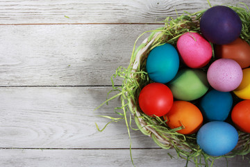 Colorful Easter eggs and pile of green grass in basket on white wooden table. Easter background, egg hunt concept. Blank copy space, mockup, hay