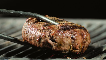 The chef puts a semi-cooked burger patty on the grill with iron tongs