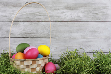 Colorful eggs in basket on green grass against white wooden wall. Easter background or egg hunt concept. Pink, yellow, green, orange eggs, hay