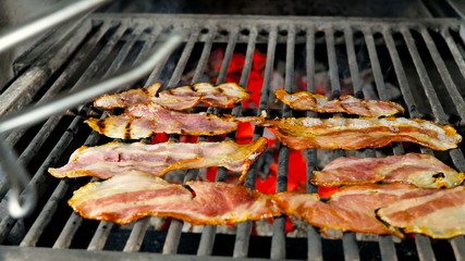 Half-cooked bacon is turned over one by one with iron tongs to the other side for final cooking on the grill