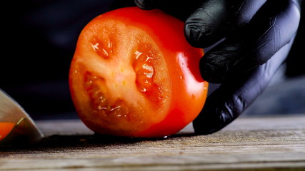 The chef of the restaurant in black gloves and with a large knife cuts red tomato on a wooden plank...