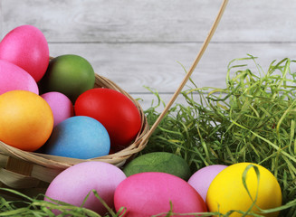 Colorful eggs scattered from basket on green grass against white wooden wall. Easter background or egg hunt concept. Pink, yellow, green, orange eggs, hay