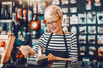Smiling Caucasian female worker with short blonde hair and eyeglasses using cash register while standing in bicycle store. - obrazy, fototapety, plakaty
