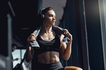 Fototapeta na wymiar Young attractive strong muscular female bodybuilder with ponytail and headphones posing in gym with towel around neck. When life is tough, remember you are tougher.