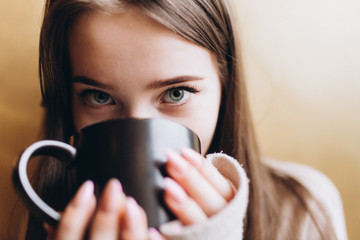 Cheerful young girl having breakfast at cozy home with big cup of coffee or tea holding in hands. Lifestyle, relaxation, natural beauty concept