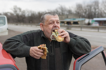Portrait of Caucasian senior driver gobbling lyulya kebab in lavash near his car