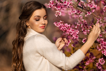 Young beautiful teen girl with perfect skin and makeup is wearing romantic clothes posing near blossom tree in cherry garden