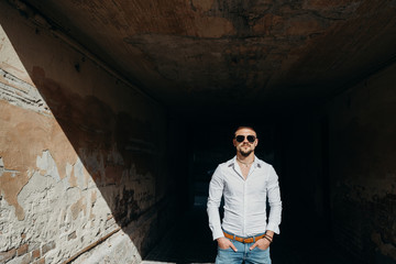 Obraz premium Colorful portrait of a handsome man dressed in white shirt and sunglasses on the grunge background. Happy young man posing outdoor, enjoying the sunny weather.