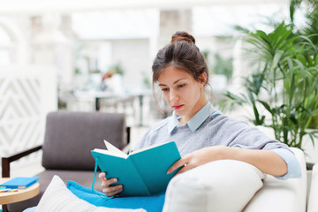 Young woman is reading book or notepad. Girl is sitting on sofa in cozy modern cafe indoors....