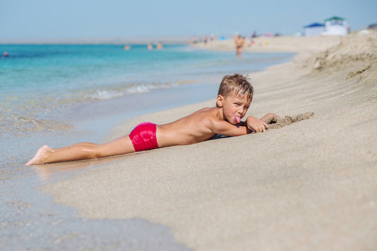 A little boy lying on the sand on the beach is dabbling and showing his tongue