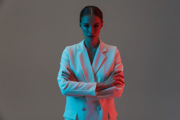 Photo of serious woman 20s wearing elegant jacket looking at camera and standing under neon lights