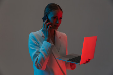 Image closeup of smart woman 20s holding smartphone and working on laptop while standing under neon...