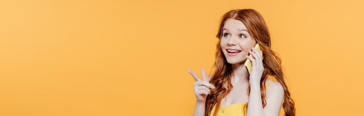 panoramic shot of beautiful smiling redhead girl pointing with finger while talking on smartphone