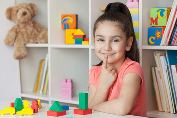 Back to school and happy time! Happy cute child girl sitting at a table with a white bookcase with colored books and toys behind. 