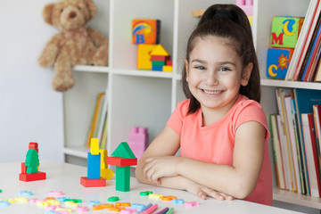 Back to school and happy time! Happy cute child girl sitting at a table with a white bookcase with colored books and toys behind. 