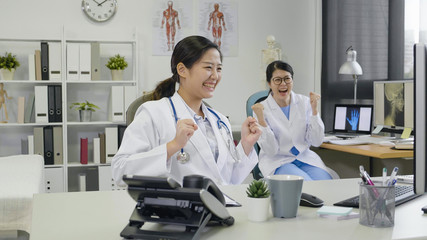Extremely happy medical professional showing winning sign. female employees joyfully with hands in fist laughing looking computer screen of good news. doctors with success report in hospital office