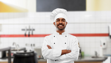 cooking, profession and people concept - happy male indian chef in toque with crossed arms over restaurant kitchen background