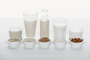 glasses and bottle with coconut, chickpea, oat, rice and almond milk on white wooden table with ingredients in bowls isolated on white