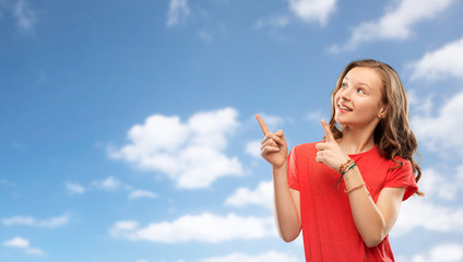 people concept - smiling teenage girl with long hair in red t-shirt pointing fingers to something over blue sky and clouds background