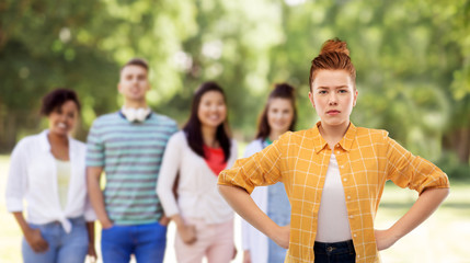 people concept - displeased red haired teenage girl in checkered shirt with hands on hips over group of friends at summer park background