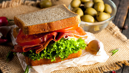 Close-up photo of a club sandwich. Sandwich with meat, prosciutto, salami, salad, vegetables, lettuce, tomato, onion and mustard on a fresh sliced rye bread on wooden background. Olives background.