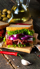 Close-up photo of a club sandwich. Sandwich with meat, prosciutto, salami, salad, vegetables, lettuce, tomato, onion and mustard on a fresh sliced rye bread on wooden background. Olives background.