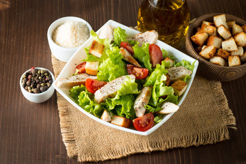 Fresh salad made of tomato, ruccola, chicken breast, eggs, arugula, crackers and spices. Caesar salad in a white, transparent bowl on wooden background