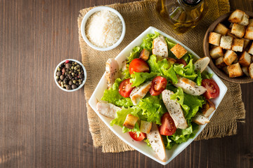 Fresh salad made of tomato, ruccola, chicken breast, eggs, arugula, crackers and spices. Caesar salad in a white, transparent bowl on wooden background