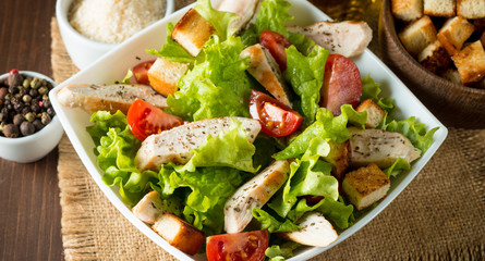 Fresh salad made of tomato, ruccola, chicken breast, eggs, arugula, crackers and spices. Caesar salad in a white, transparent bowl on wooden background