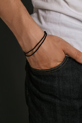 Cropped closeup shot of man's hand, wearing black lucky rope bracelet with 2 rows of thick string. The guy is wearing jeans and shirt, putting his hand into the pocket, posing on dark background.