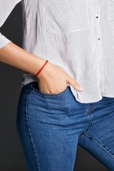 Cropped half-turn shot of woman's hand, wearing red lucky rope bracelet with fishtail plaiting. The lady is wearing jeans and stripy shirt, putting her hand into pocket, posing on dark background.