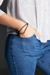 Cropped half-turn shot of woman's hand, wearing 2-row black lucky rope bracelet with milky bead. The lady is wearing jeans and stripy shirt, putting her hand into pocket, posing on dark background.