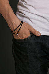 Cropped half-turn shot of man's hand, wearing 2-row black lucky rope bracelet with milky bead. The guy is wearing black jeans and t-shirt, putting his hand into pocket, posing on the dark background.