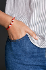 Cropped closeup shot of lady's hand, wearing red lucky rope bracelet, adorned with 3 milky beads. The girl is wearing jeans and stripy shirt, putting hand into pocket, posing on dark background.