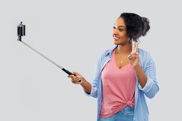 technology and people concept - happy african american woman taking picture by smartphone on selfie stick and showing peace hand sign over grey background