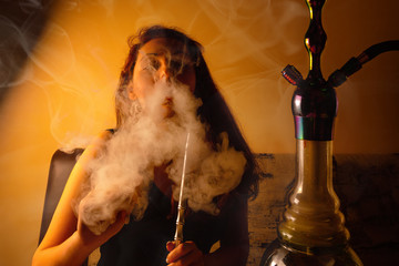Relaxing on the couch, the woman smokes a hookah and releases a cloud of steam. Around clouds of...