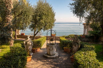 Fototapeta premium View of Lake Garda from a garden with an ancient stone well and statues in springtime, Sirmione, Lombardy, Italy