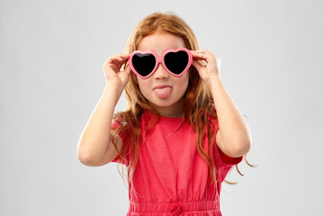 childhood, valentines day and summer concept - naughty red haired girl with heart shaped sunglasses showing tongue over grey background