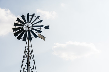 Low angle view of an old-fashioned, multi-bladed, metal wind pump atop a lattice tower in backlight against a pale blue sky with white clouds.