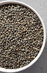 Full bowl of dry green french lentils on a gray surface, top view. Overhead, from above, flat lay.