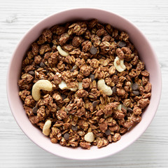 Homemade chocolate granola with nuts in a pink bowl over white wooden surface, top view. From above, overhead.