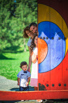 Happy Family Looks Out From Behind The Door. Mom, Dad, Daughter And Little Son For A Walk In The Park. Smiling Parents Play Hide-and-seek With Children.