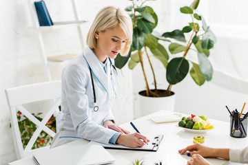 attractive blonde nutritionist in white coat writing diagnosis near patient