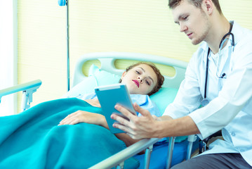 Young friendly caucasian pediatrician male doctor is examining patient woman with tablet pc on bed in hospital, consultation with stethoscope in clinic office. Healthcare and medical concept