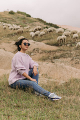 Naklejka na ściany i meble Happy young woman in a sweater against herds of sheep in the mountains