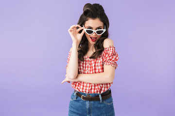 Portrait of flirting pin-up woman 20s in american style wear holding retro sunglasses and smiling...